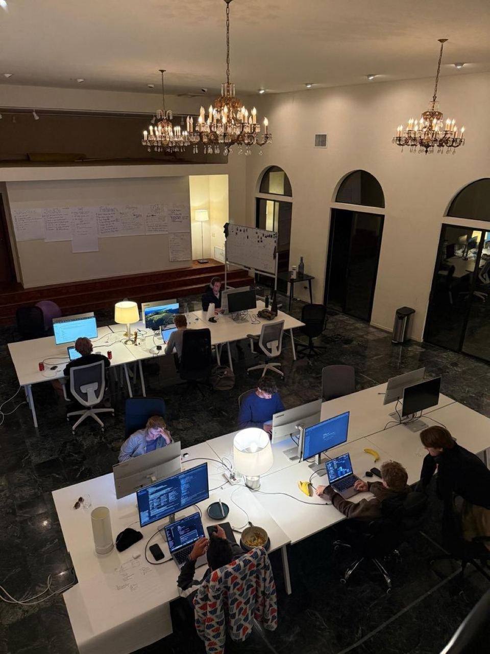 Fellows at long white desks under chandeliers in the main hall of the AI hacker house in Palo Alto, whiteboards on the stage, 2026