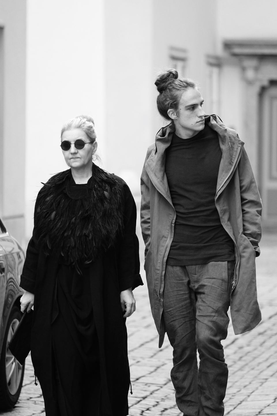 Joël Hainzl with long hair in a bun walking down a cobbled street next to a woman in a feather coat, black and white street photo from his modeling years