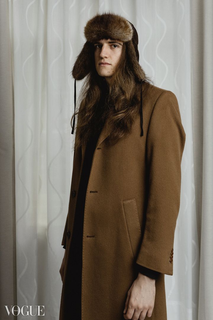 Joël Hainzl with long hair in a fur trapper hat and a long camel coat, portrait by Evgenia Rieger published on PhotoVogue, Vogue Italia, with the Vogue watermark