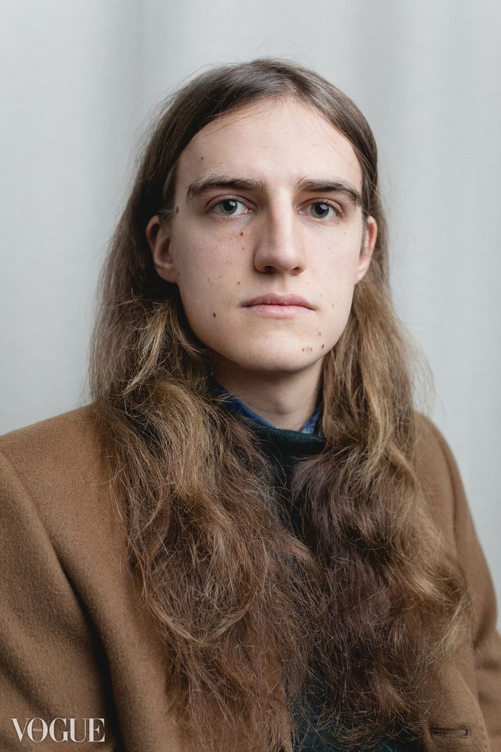 Joël Hainzl with long hair in a camel coat, studio portrait by Evgenia Rieger published on PhotoVogue, Vogue Italia, with the Vogue watermark