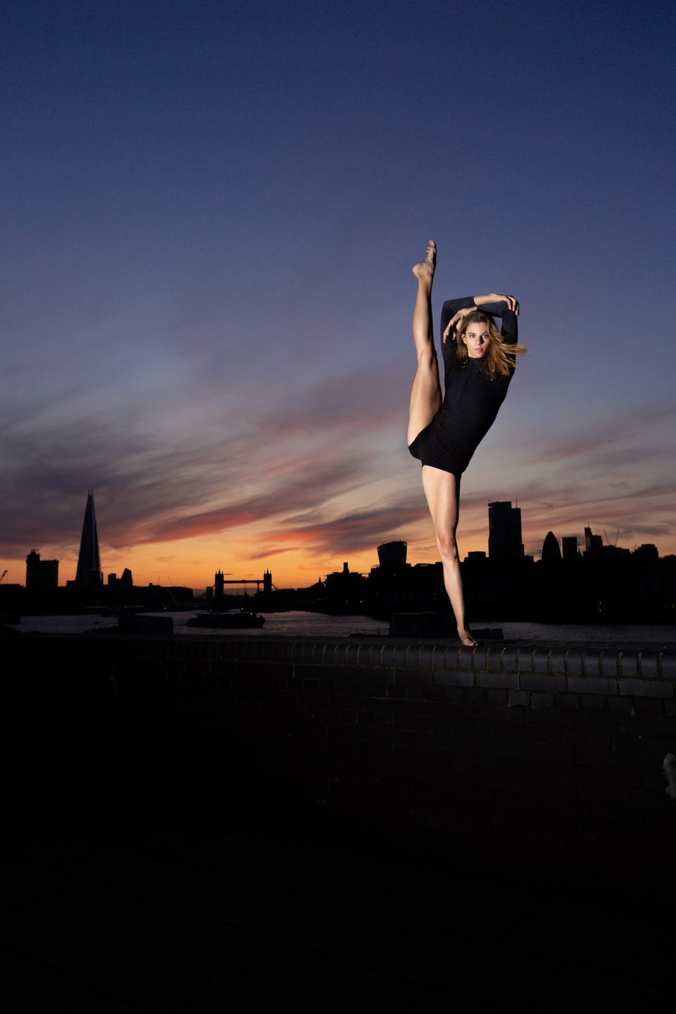 Dancer in a black leotard balancing on one leg on a brick wall at dusk, the London skyline with the Shard and Tower Bridge behind her