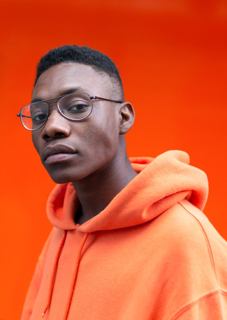 Young man in aviator glasses and an orange hoodie against an orange wall, head turned over his shoulder, studio portrait