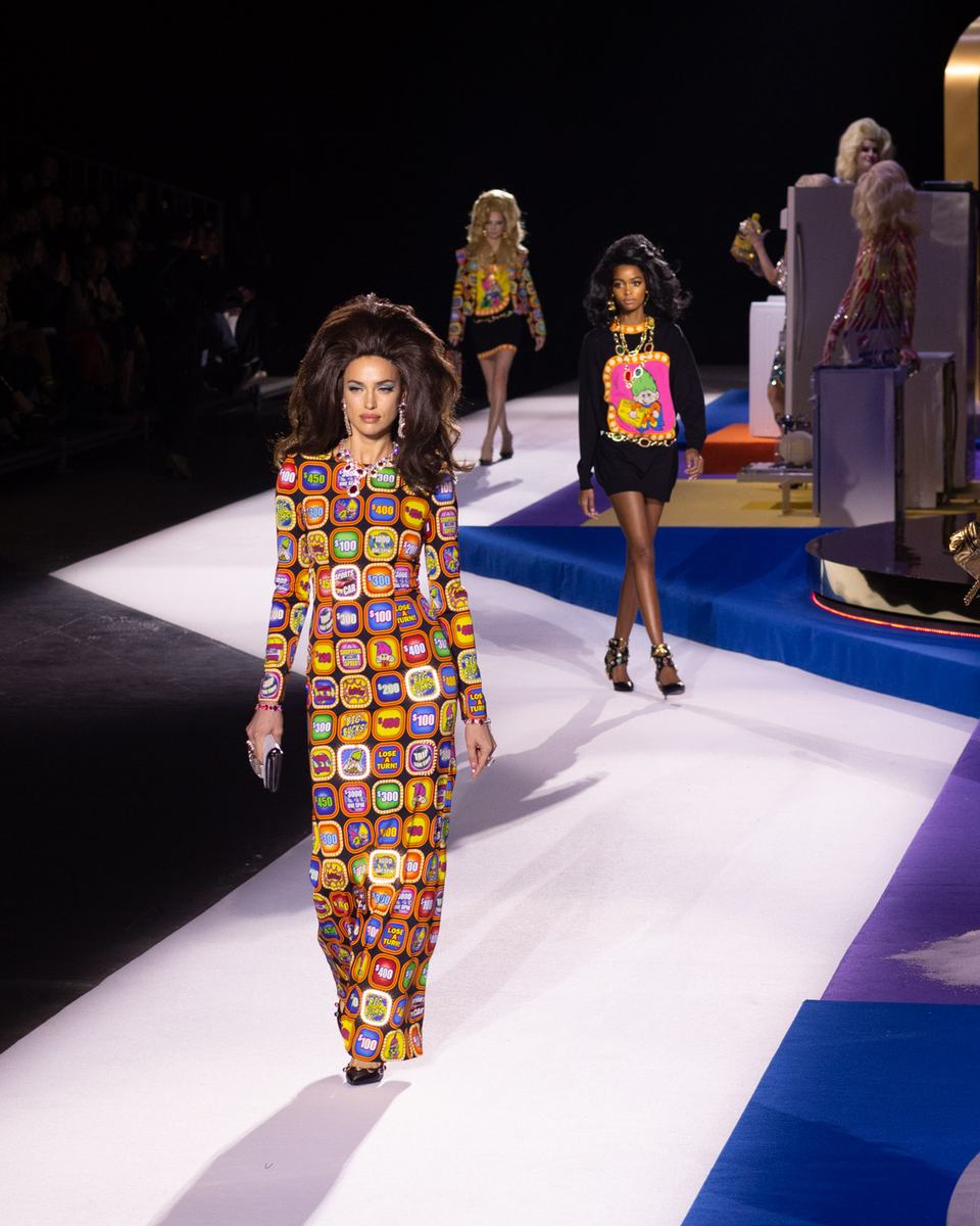 Models walking a runway at a fashion show, the lead model in a long printed gown with teased hair, more looks following behind her