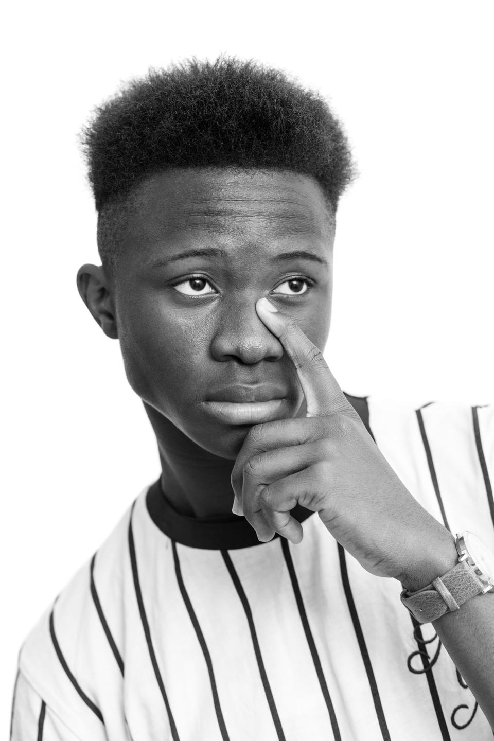 Black and white studio portrait of a young man in a pinstripe shirt, finger resting on his nose, looking off to the side