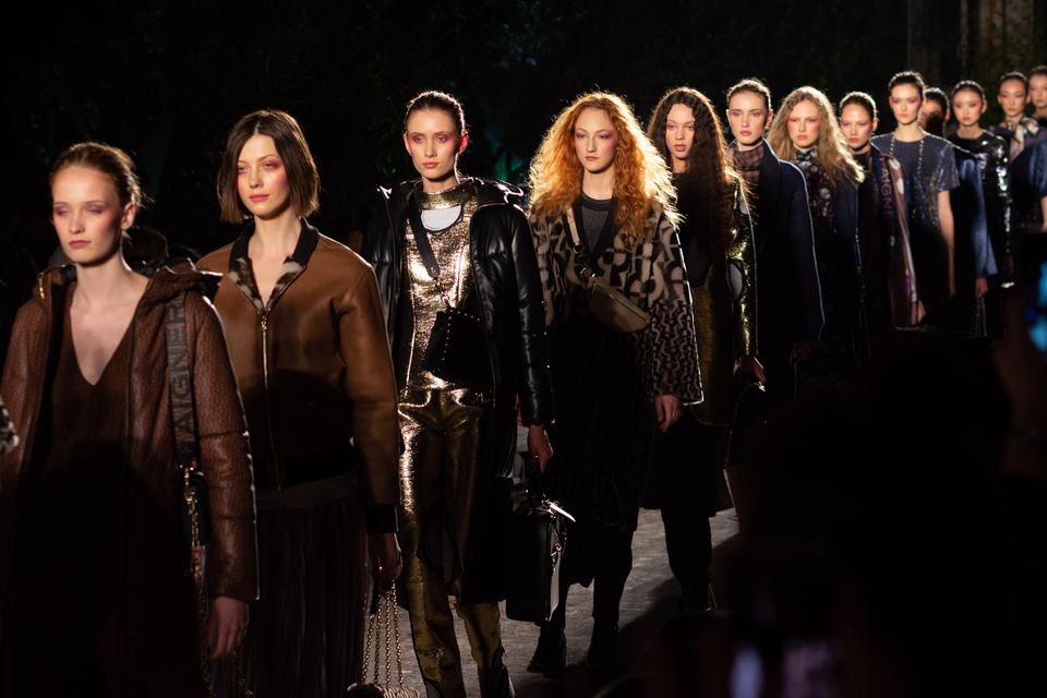 Runway finale at an evening outdoor fashion show, a long line of models in leather and shearling coats walking toward the camera