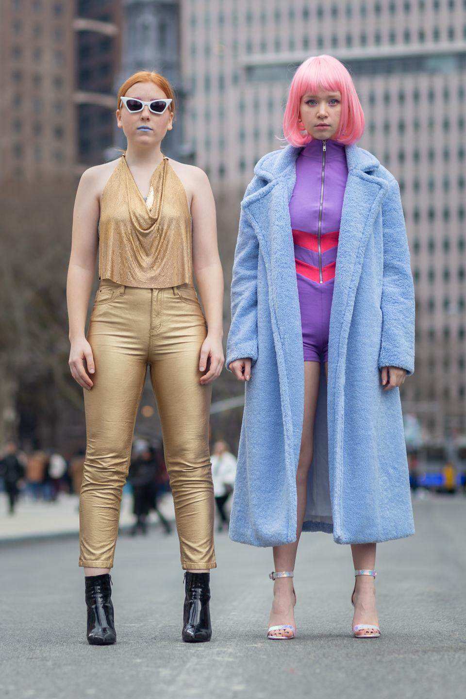 Two women in street style looks on a city plaza, one in gold with cat eye sunglasses, the other in a pink wig and a long blue coat
