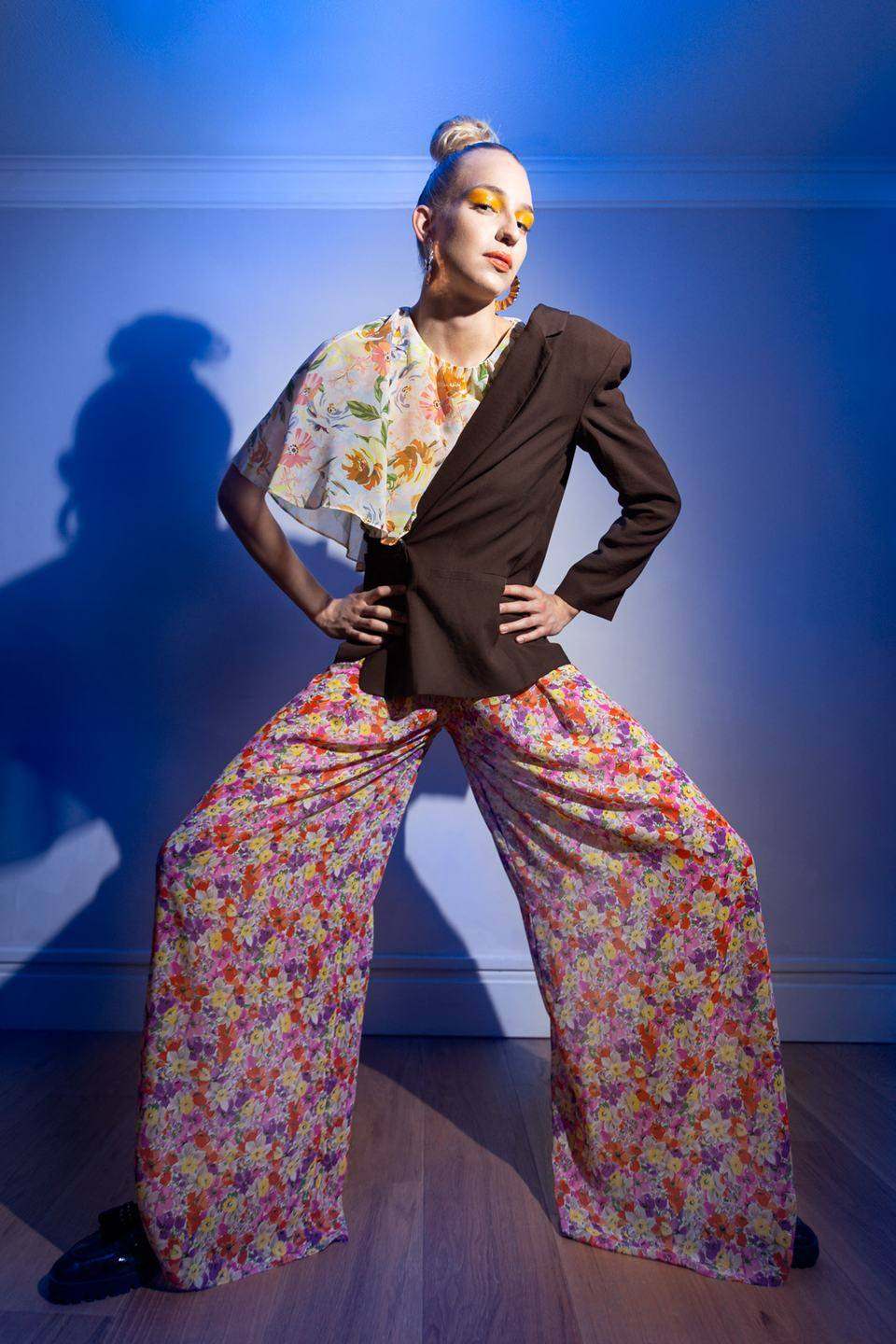 Model in wide floral trousers, a brown half blazer and a floral blouse, hands on hips, lit against a blue wall with her shadow behind her