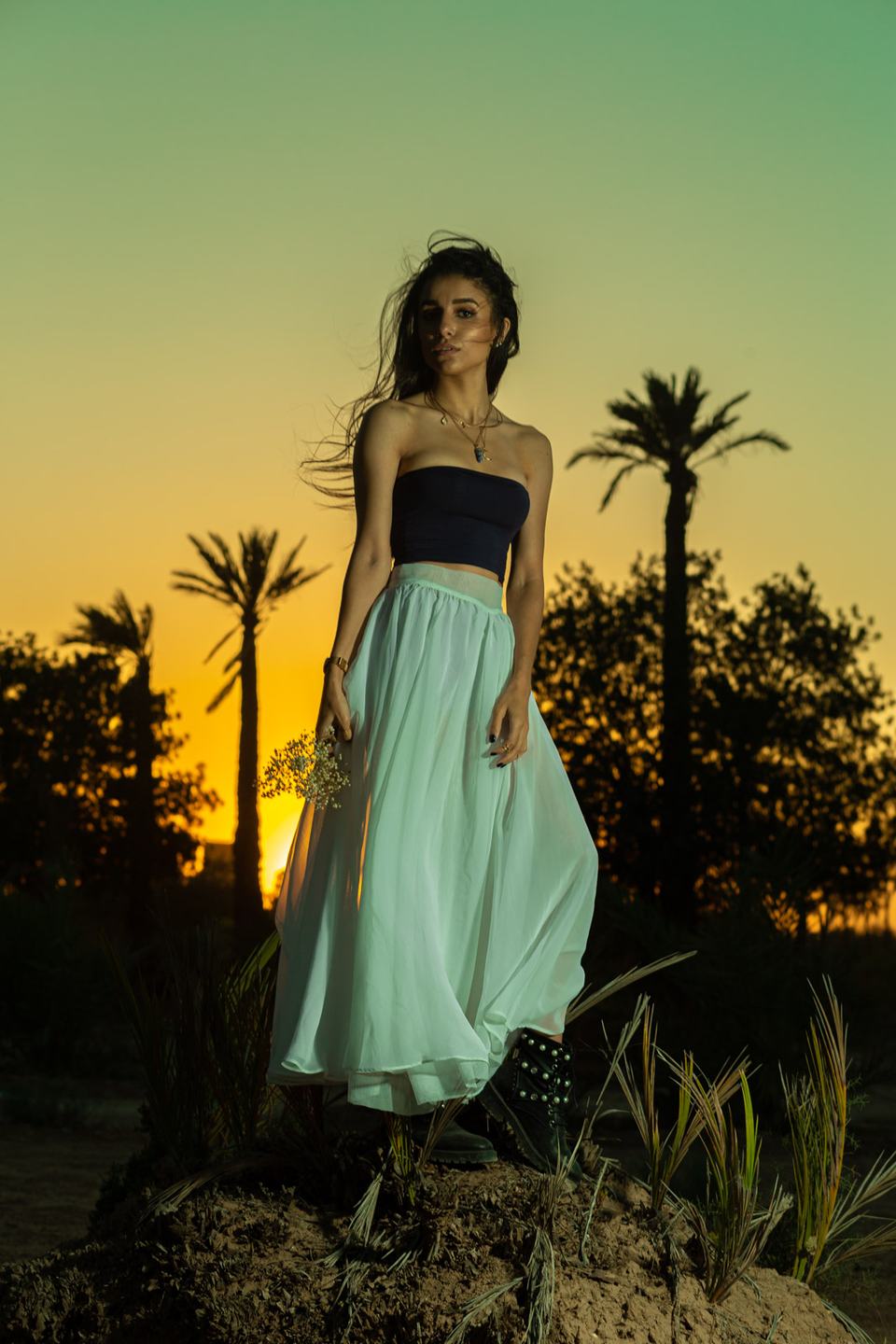 Woman in a long white skirt and dark top standing on a mound at sunset, palm trees silhouetted against a golden sky