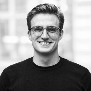 Joël Hainzl, black and white headshot, smiling in clear round glasses and a black crew neck sweater