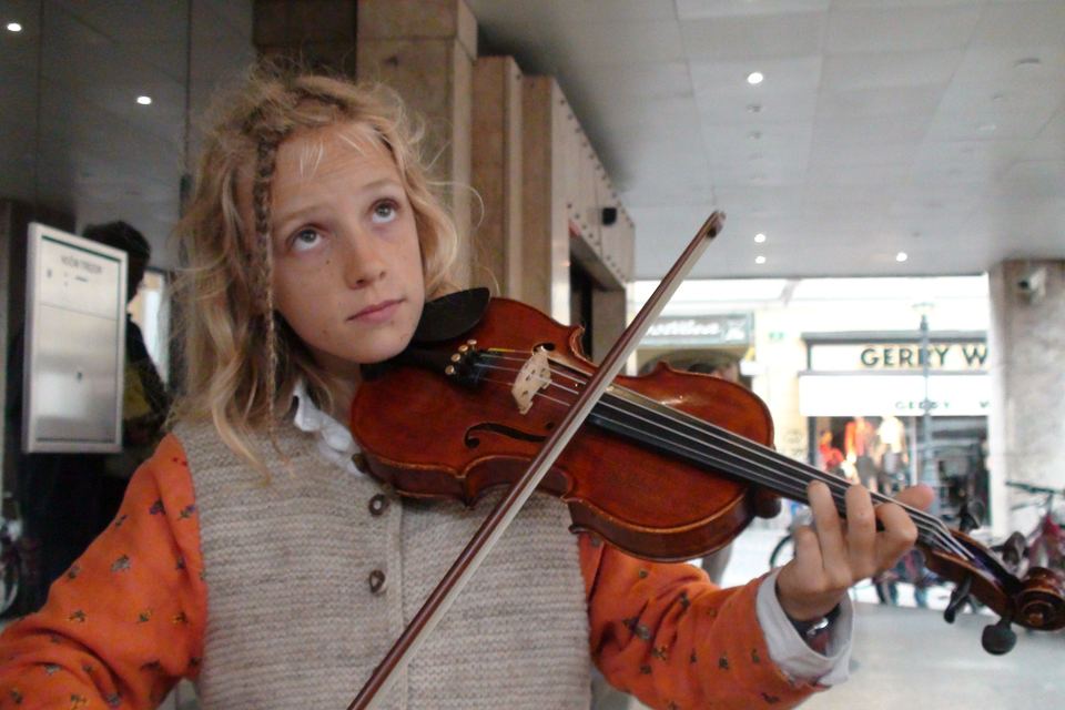 Joël Hainzl, about eight years old with curly blond hair, playing violin in a shopping arcade in Graz, bicycles and a Gerry Weber shop behind him