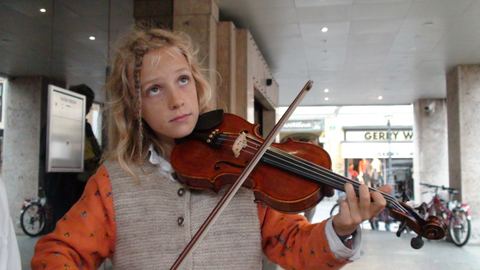 A blond child with braided hair playing the violin in a street arcade, bicycles and shop fronts behind