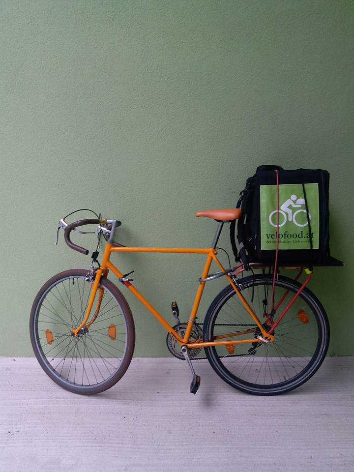Joël Hainzl's orange road bike with the velofood.at delivery box on the rack, against a green wall in Graz